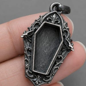 Puede incluir: Colgante plateado con un centro en forma de ata&uacute;d. El colgante tiene un marco ornamentado de estilo g&oacute;tico con volutas y elementos decorativos. La argolla es un simple lazo para una cadena. El dise&ntilde;o general es oscuro y detallado.