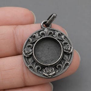 Op de afbeelding: Zilverkleurige hanger met een cirkelvormig ontwerp. De hanger heeft een centraal verzonken gebied omgeven door een sierlijk bloemmotief met rozen en krullende bladeren. Bovenop zit een kleine lus voor een ketting.