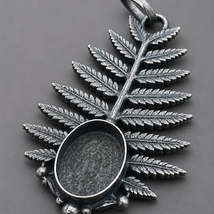 May include: A dark silver-toned metal pendant featuring a detailed fern leaf design with a central oval-shaped bezel setting. The surface has an oxidised, textured finish with small spherical accents at the base and a sturdy loop at the top.