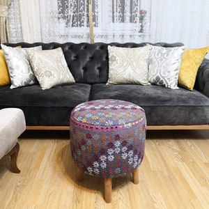 Puede incluir: Un sofá gris capitoné con una variedad de almohadas decorativas con patrones, incluyendo dorado, plateado y crema. Un puf con patrones coloridos se encuentra frente al sofá sobre un suelo de madera.