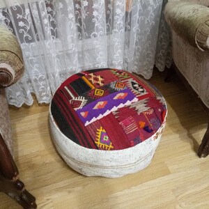 May include: A round, white and cream colored ottoman with a colorful, patterned fabric top. The fabric is a patchwork of red, purple, black, and yellow geometric designs.