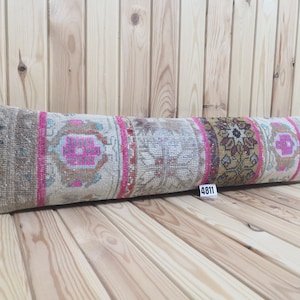 Pode incluir: Uma almofada longa e retangular com fundo bege e padrões geométricos coloridos em rosa, marrom e dourado. A almofada é feita de tecido tecido e tem uma borda franjada. A almofada está sobre uma superfície de madeira.