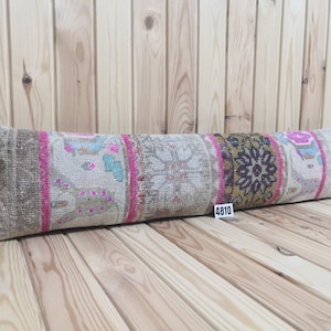 Op de afbeelding: Een lang, rechthoekig kussen met een kleurrijke, gepatenteerde stof. Het kussen is gemaakt van een patchwork van verschillende stoffen, elk met een uniek ontwerp. Het kussen is beige, bruin, roze en blauw. Het kussen ligt op een houten oppervlak.