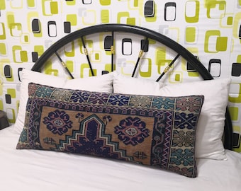 May include: A long, rectangular pillow with a colorful, patterned design. The pillow is made of a woven fabric with a brown background and intricate blue, purple, and green patterns. The pillow is sitting on a white bed with a black metal headboard.