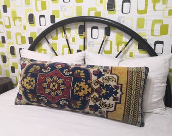 May include: A long, rectangular throw pillow with a colorful, intricate pattern in shades of red, blue, yellow, and white. The pillow is made of a woven fabric and has a fringed edge.