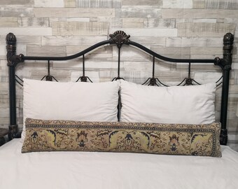 May include: A decorative bolster pillow with a floral pattern in shades of beige, brown, and blue. The pillow is placed on a white bed with a black metal headboard.