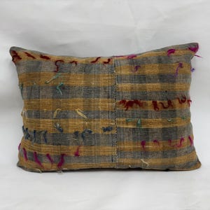 Puede incluir: Cojín rectangular con un patrón de rayas en tonos grises y dorados. El cojín tiene acentos coloridos y texturizados en rojo, rosa y verde azulado. El cojín es un elemento decorativo para el hogar.
