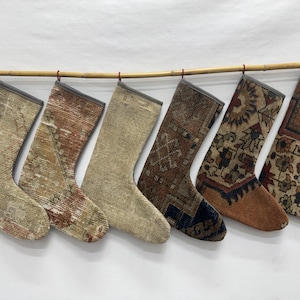 Puede incluir: Seis calcetines navideños colgados en fila de una vara de bambú. Cada calcetín está confeccionado con material de alfombra vintage que presenta patrones tejidos distintos y paletas de colores que incluyen tonos beige, marrón, óxido y azul con borde gris.