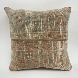 May include: Square cushion with a woven texture. The cushion features a colour palette of muted greens, pinks, and browns, with a horizontal brown band across the centre. The design includes a grid-like pattern.