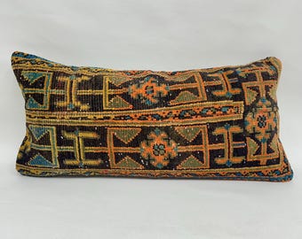 May include: Rectangular pillow with a geometric pattern in shades of brown, orange, green, and blue. The pillow has a textured appearance, suggesting a woven or embroidered design. The pattern features repeating shapes and floral motifs.