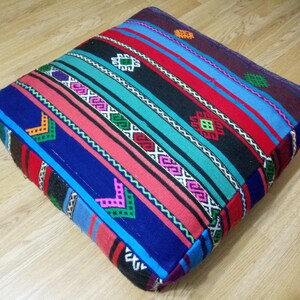 May include: A colorful square floor cushion with a striped pattern in shades of blue, red, pink, green, and black. The cushion has a geometric design with intricate details.