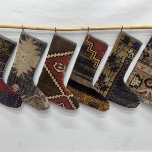 May include: Six decorative stockings hanging in a row from a bamboo rod. Each stocking features a unique woven pattern with varied color palettes including deep red, navy blue, brown, beige, and charcoal gray, finished with a simple gray trim.