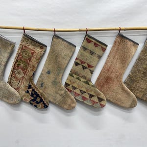 May include: Six holiday stockings hanging from a bamboo rod. Each stocking features a unique textile pattern, including floral motifs, geometric shapes, and distressed textures in muted tones of beige, brown, red, blue, and cream with grey trim.