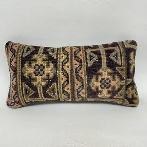 May include: Rectangular decorative pillow with a geometric pattern. The pillow features a dark brown background with a repeating design in shades of green, tan, and pink. The pillow is approximately 20 inches long.