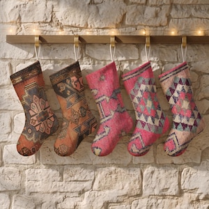 May include: Five holiday stockings hanging from a wooden rack, each featuring a unique woven pattern. The stockings display a variety of colours including orange, brown, pink, teal, and cream, with intricate geometric and floral-inspired designs.