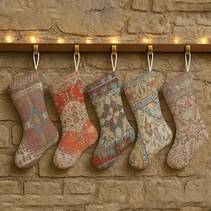 May include: Five Christmas stockings with ornate, colorful patterns hang from a wooden beam. The stockings feature designs in shades of red, orange, blue, and beige, with a vintage, rug-like aesthetic. A string of warm white lights is strung above.
