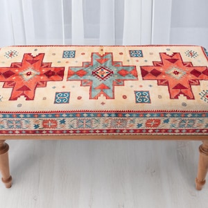 May include: A rectangular bench with a beige upholstered top featuring a geometric pattern in red, teal, and blue. The bench has a wooden frame and four turned legs, suitable for a living room or entryway.