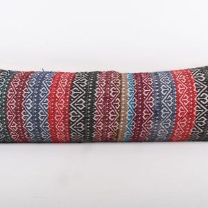 May include: A long, rectangular pillow with a colorful, patterned design. The pillow features vertical stripes in red, blue, green, and black, with a repeating white geometric pattern. The pillow is a decorative home accent.