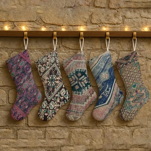 May include: Five Christmas stockings with patterned designs hang from a wooden rod with gold hooks. The stockings feature various colors, including blue, pink, and brown, and are suspended against a stone wall with string lights.