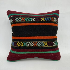 May include: A colorful striped pillow with a geometric pattern. The pillow is made of woven fabric and has a black background with orange, red, and green stripes. The geometric pattern is in shades of blue, yellow, and white.