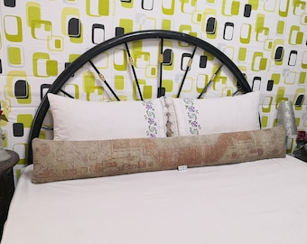 May include: A long, rectangular pillow with a faded, patterned design in shades of brown, beige, and red. The pillow is placed on a white bed, with a white pillow behind it. The headboard is black.