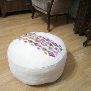May include: White round pouf with colorful geometric embroidery. The pouf is made of a soft, textured fabric and has a round shape.