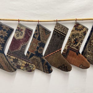 Op de afbeelding: Zes kerstsokken hangen op een rij, elk gemaakt van vintage tapijtfragmenten met verschillende geometrische en bloemmotieven in diverse kleurenpaletten, waaronder marineblauw, donkerrood, bruin, oranje en crème met grijze randen.