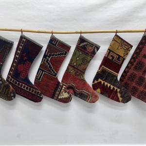 May include: Six Christmas stockings hanging in a row on a bamboo rod. Each stocking features a unique, intricate woven pattern with varied color palettes including deep reds, blues, greens, yellows, and dark browns, resembling vintage rug textures.