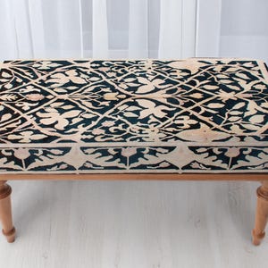 May include: A rectangular ottoman with a dark teal and cream floral pattern on the top. The ottoman has a wooden frame and four turned legs. The fabric design features a repeating pattern of stylized leaves and vines.