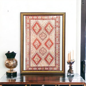 Puede incluir: Una alfombra vintage enmarcada con un patrón geométrico rojo, blanco y dorado. La alfombra se exhibe sobre una superficie de madera oscura con un candelero decorativo y un jarrón.