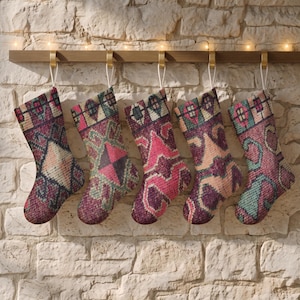 Könnte beinhalten: Fünf Weihnachtssocken hängen in einer Reihe, jede mit einem einzigartigen Webmuster in den Farben tiefes Magenta, Blaugrün und Creme. Die Socken sind mit weißen Schlaufen an einer Holzleiste befestigt und zeigen komplexe geometrische Muster.