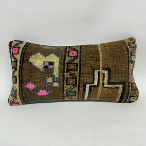 May include: Rectangular brown pillow with a woven texture and geometric patterns. The design includes pink, beige, and black accents. The pillow has a vintage or antique aesthetic, suitable for home decor.