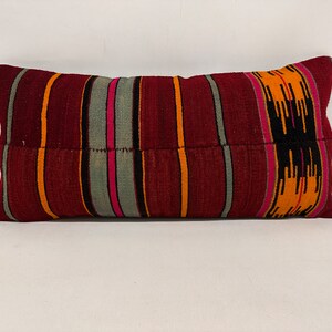 May include: A long, rectangular pillow with a striped pattern in shades of red, orange, black, gray, and pink. The pillow is made of a woven fabric and has a slightly textured surface.