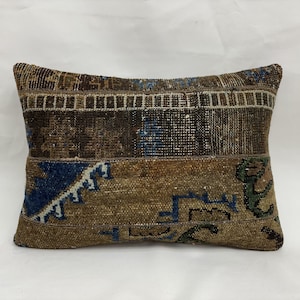 May include: Rectangular decorative pillow with a patchwork design. The pillow features a mix of brown, blue, and green hues. The design includes geometric shapes and patterns, creating a vintage or rustic aesthetic. The pillow is likely made of a woven fabric.