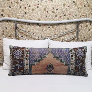 May include: A long, rectangular pillow with a colorful, patterned fabric cover. The pillow is on a white bed with a white headboard. The headboard has a decorative metal design.