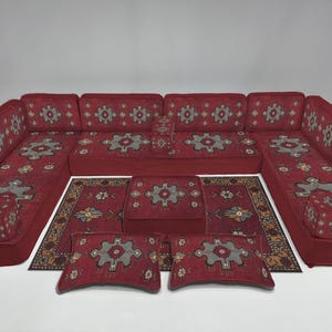 Puede incluir: Un juego de sofás modulares rojos con cojines estampados y alfombras a juego. El conjunto incluye un gran sofá seccional, una otomana cuadrada, dos cojines rectangulares y varias alfombras pequeñas. Los cojines y las alfombras presentan un diseño floral gris y beige.