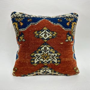May include: Square decorative pillow with a vintage rug design. The pillow features a rust-colored center with geometric patterns in white and yellow. The border is a deep blue with additional geometric details.