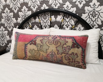 May include: A rectangular decorative pillow with a vintage rug design. The pillow features a pink border, a beige center with green and blue patterns, and is placed on a white bed with a patterned headboard.