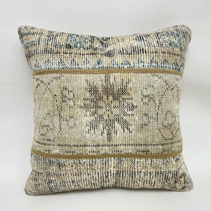May include: Square decorative pillow with a vintage, distressed look. The pillow features a central floral design in beige and brown, framed by a tan border. The top and bottom sections have a striped pattern with blue and beige accents.