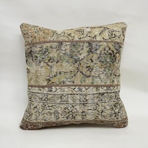May include: Square decorative pillow with a vintage rug design. The pillow features a beige, brown, and green color palette with intricate patterns. The pillow has a rectangular shape and is likely used for home decor.