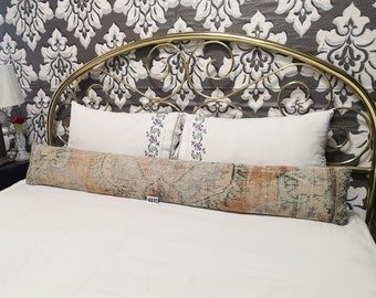 May include: A long, rectangular, patterned pillow with a beige, orange, and brown color scheme. The pillow is on a white bed with a gold metal headboard. The headboard has a floral pattern.
