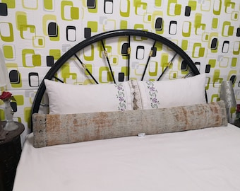 May include: A long, rectangular, gray and brown patterned bolster pillow on a white bed with a black metal headboard. The headboard has a geometric pattern of green and black squares on a white background. The pillow has a tag with the number 2212 on it.
