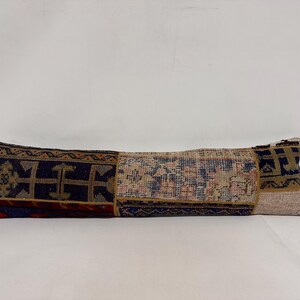 May include: A long, rectangular cushion with a patchwork design. The cushion is made of different pieces of fabric with various patterns and colours, including blue, red, brown, and beige.