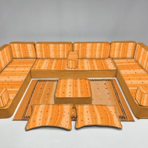 Puede incluir: Un juego de sofás modular a rayas naranjas y amarillas, con cojines a juego y una pequeña alfombra. El conjunto incluye múltiples secciones para arreglos flexibles. Los cojines y la alfombra tienen un patrón de puntos.