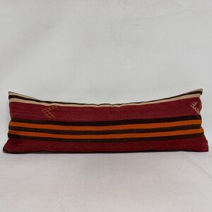 May include: A long, rectangular pillow with a red, orange, brown, and white striped pattern. The pillow has a white background with embroidered details in the corners.