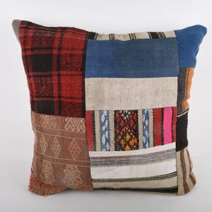 May include: A square patchwork cushion with a variety of fabric patterns and colours. The cushion features sections of red and black plaid, blue, beige, and patterned textiles. The cushion is a decorative item.