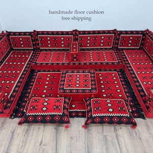 Puede incluir: Un juego de cojines de suelo hechos a mano en rojo y negro. El juego incluye varios cojines de suelo, una alfombra grande y dos almohadas más pequeñas con borlas. Los cojines y la alfombra presentan un patrón geométrico.