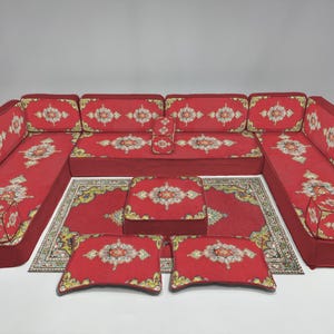 Op de afbeelding: Een rode hoekbank met een bijpassend tapijt en kussens. De bank en kussens hebben een gedetailleerd bloemmotief in wit, geel en groen. Het tapijt heeft een vergelijkbaar ontwerp. De set is op een wit oppervlak geplaatst.