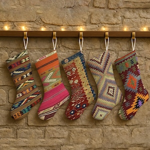 May include: Five festive Christmas stockings with detailed woven designs are suspended from a wooden rail against a stone wall. The stockings display a range of colours, including red, orange, blue, and green, and are hung by white loops.