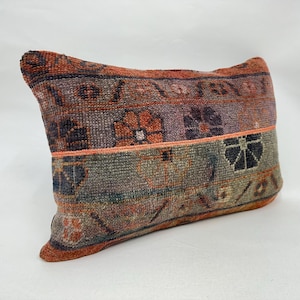 Puede incluir: Cojín decorativo rectangular con un aspecto vintage y desgastado. El cojín presenta un diseño de patchwork en tonos naranja, morado y verde. Tiene una superficie texturizada y una banda de color coral en el medio.